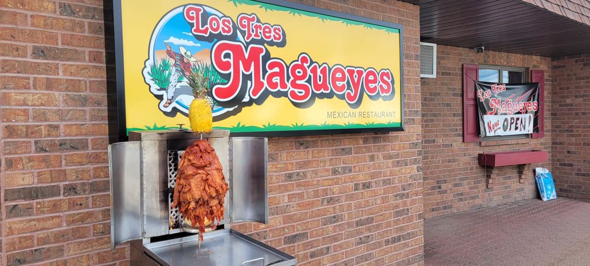 A vertical al pastor spit, loaded with meat and topped with a pineapple, standing under a sign reading Los Tres Magueyes Mexican Restaurant.