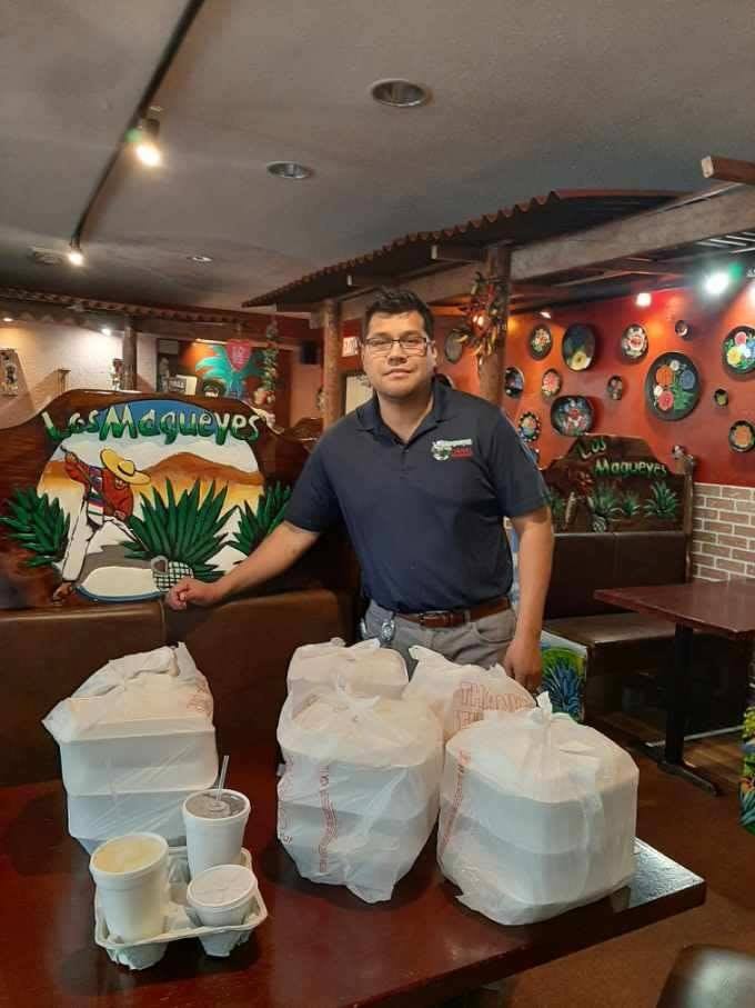 A member of staff standing behind four large bagged takeaway orders and a tray of drinks, in a dining room with hand-painted booths.