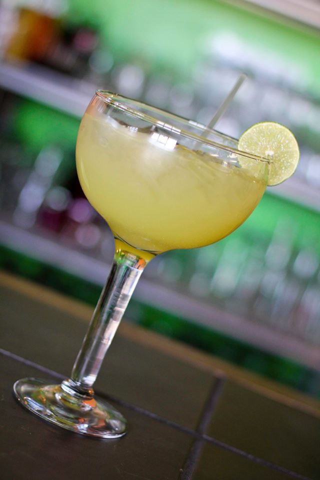 A margarita in a coupe glass with a lime wheel, on the bar in front of a lit backbar.