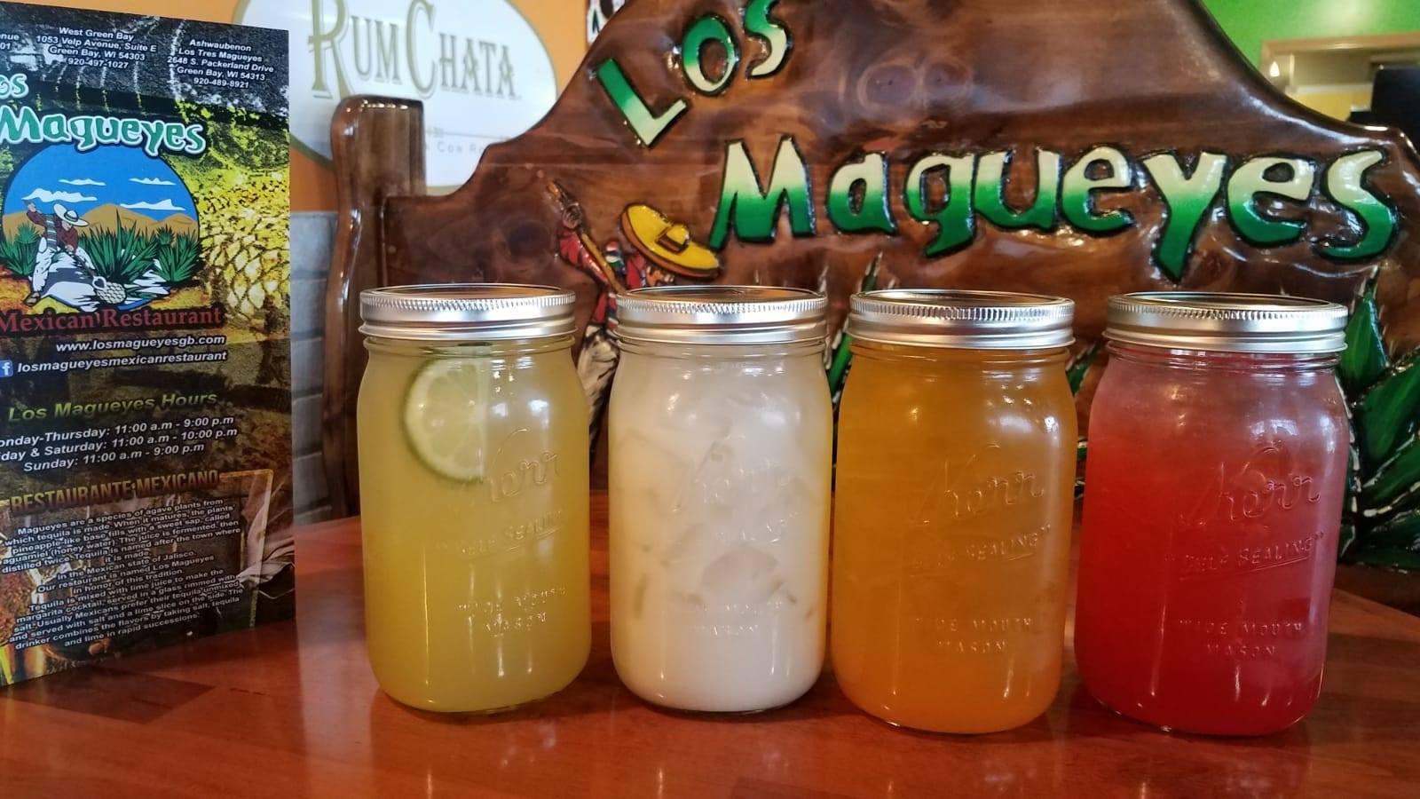 Four margaritas sealed in mason jars — lime, horchata, mango and strawberry — in front of a carved wooden Los Magueyes sign.