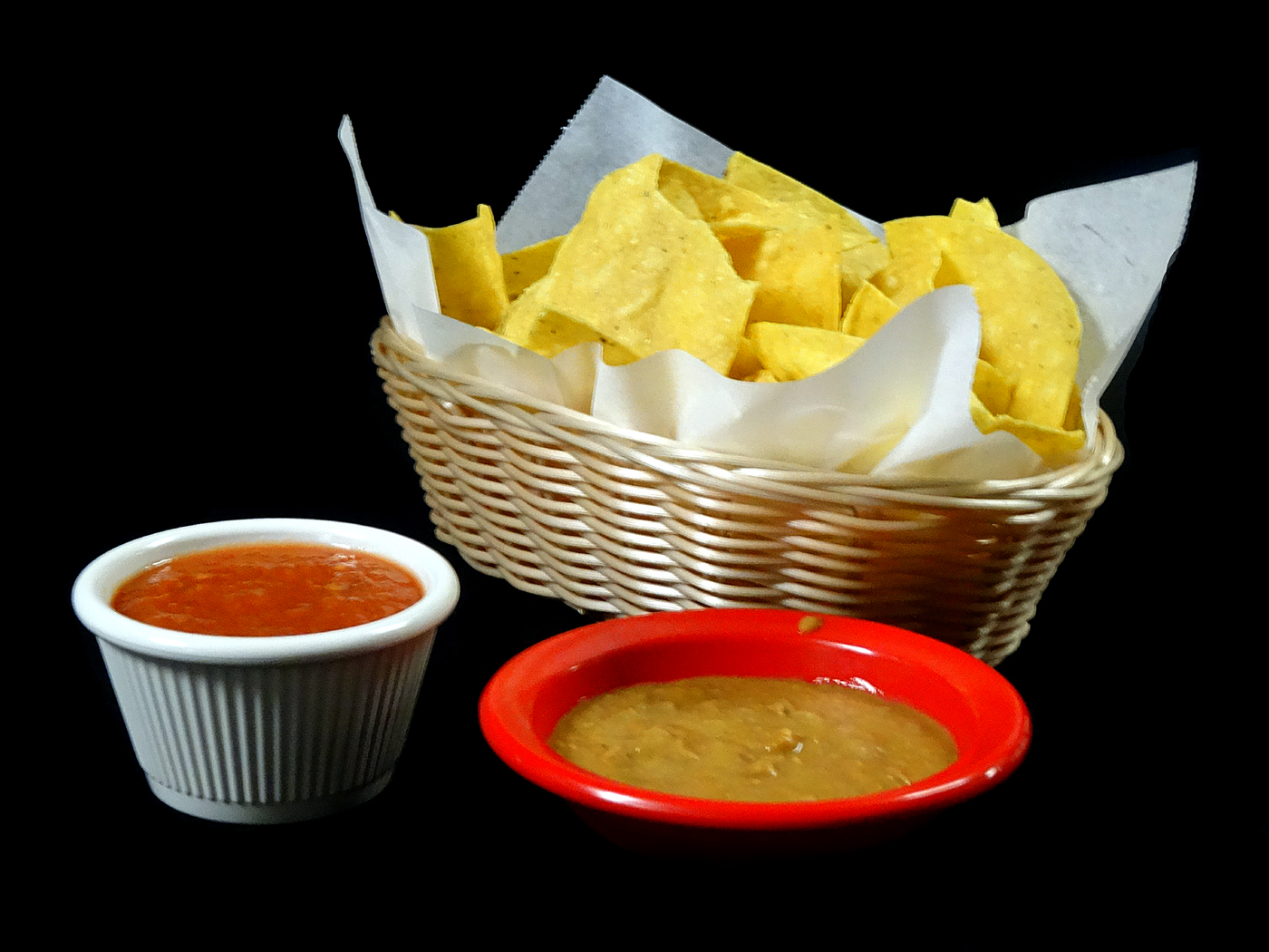 A basket of tortilla chips with a ramekin of red salsa and a red dish of bean dip.