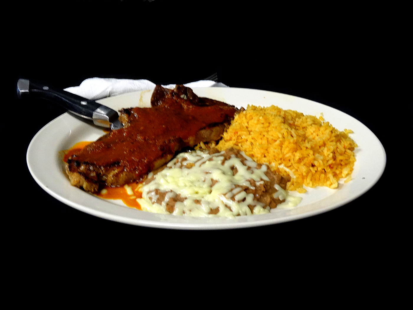 A steak under red ranchera sauce with refried beans and Spanish rice, a steak knife resting on the plate.