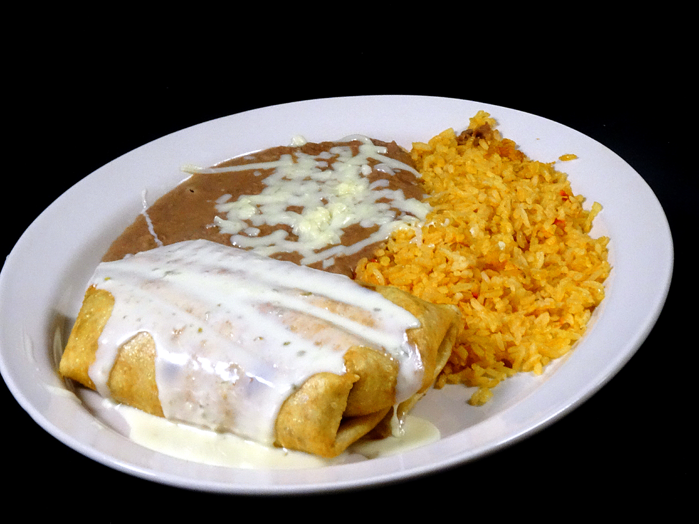 A chimichanga under white cheese dip, served with refried beans topped with melted cheese and a mound of Spanish rice.
